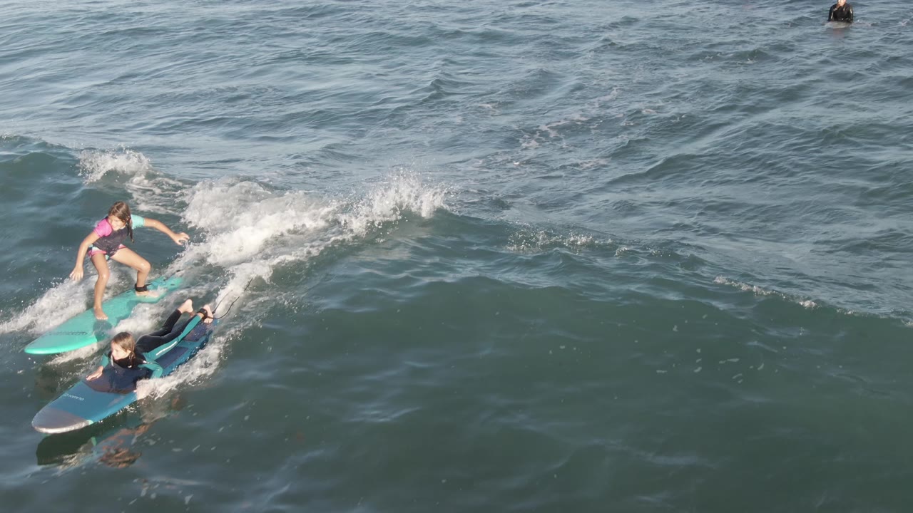 At Costa Azul Los Cabos, the aerial view places the child on the turquoise board above the instructor on a dark board, with white foam spreading across the wave face.