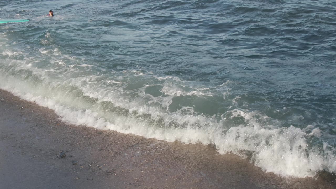 At Costa Azul Los Cabos, a person floats near the top of the frame while white surf breaks over shallow blue green water beside a dark sandy shoreline.