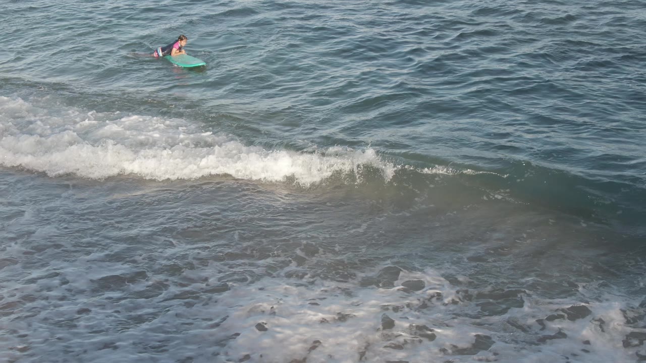 At Costa Azul Los Cabos, a person on a green bodyboard floats beyond a white breaking wave with wet sand and foam below.