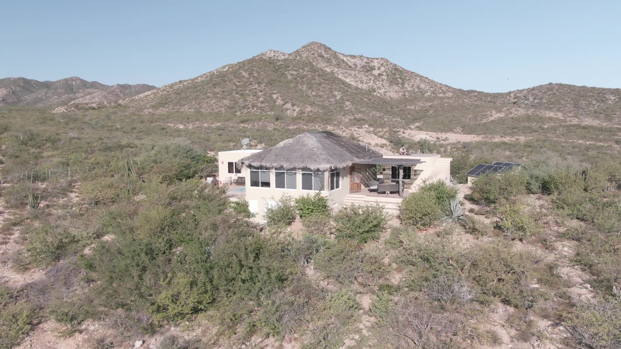 Wide elevated view in Cabo Pulmo, Baja California Sur, showing a light-colored single-story house amid shrubs and cacti with rocky mountains behind it and no visible people.