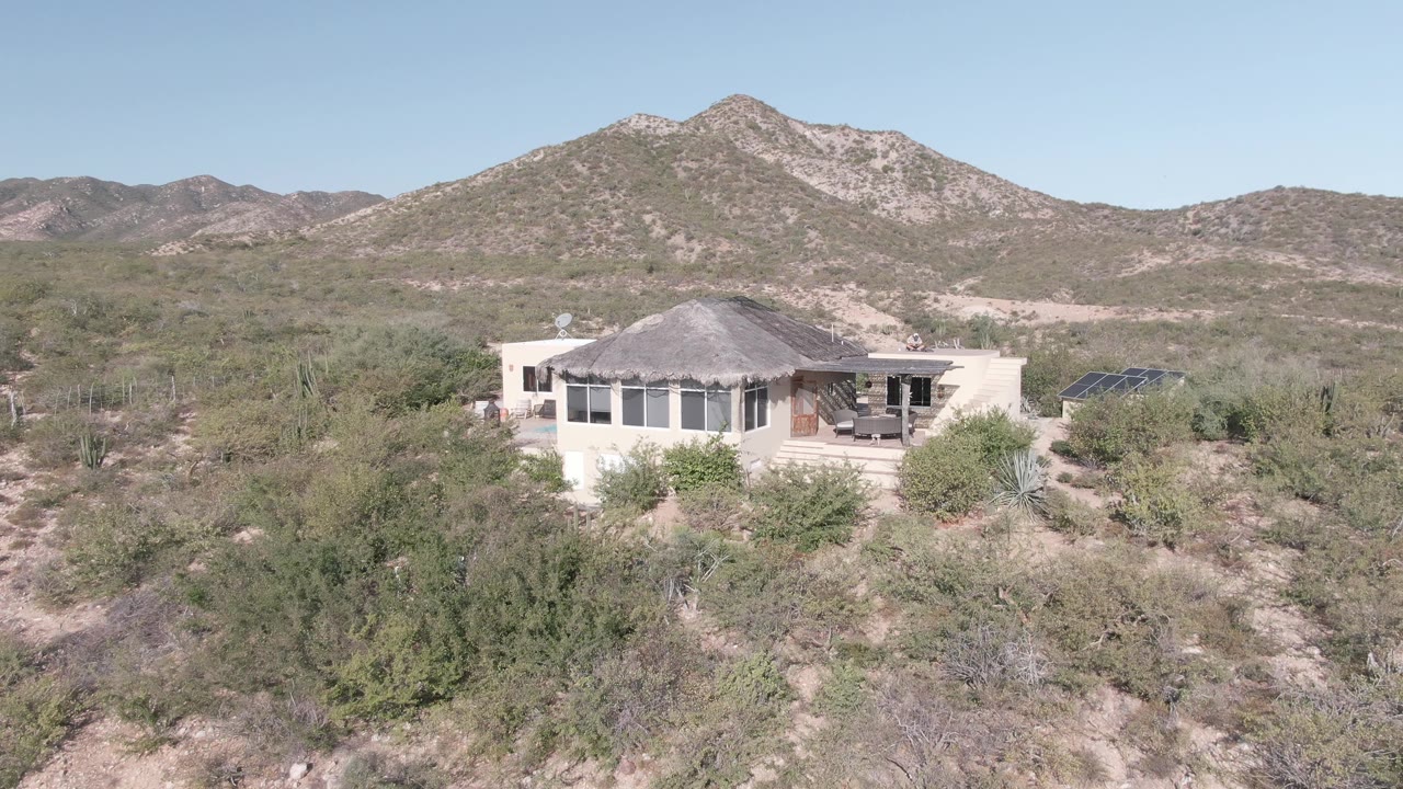 Cabo Pulmo frame with the house slightly left of center, low desert shrubs in the foreground, scattered cacti around the property, pale mountain slopes in back, and no visible people.