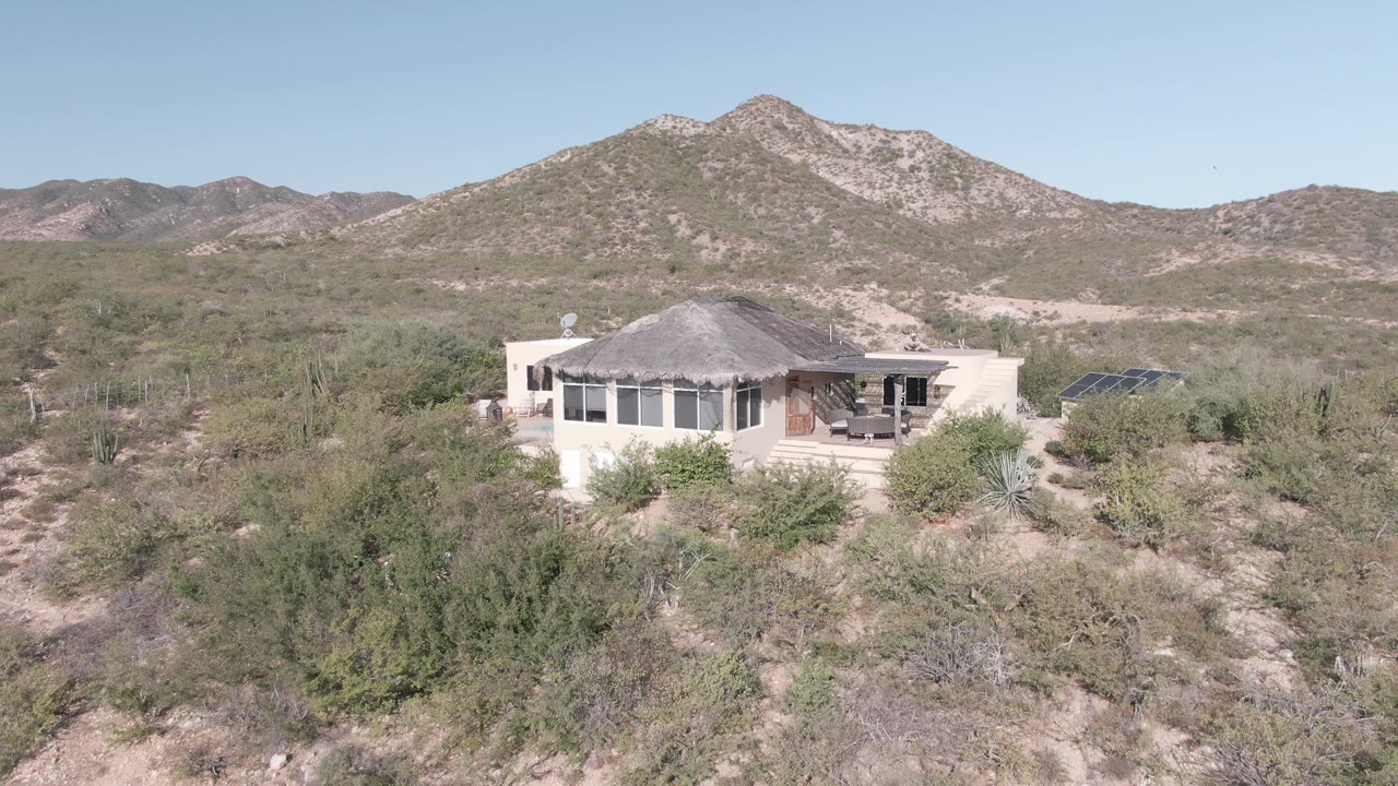 Elevated Cabo Pulmo view of a light-colored desert home with a gray roof, dark window openings, scrub vegetation, cacti, low mountains, and no visible people.