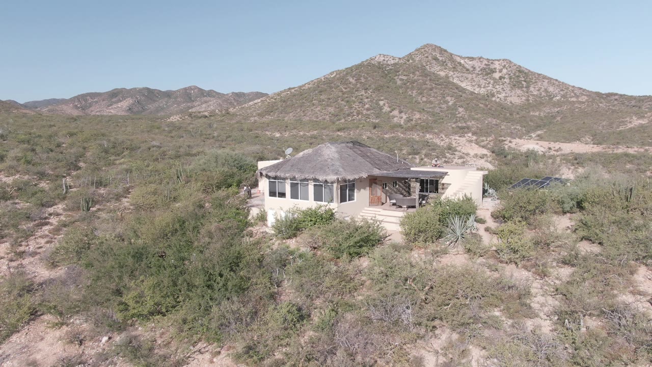 Elevated view of a light-colored house in Cabo Pulmo desert scrub, with cactus, sandy ground, low mountains, clear sky, and no visible people.