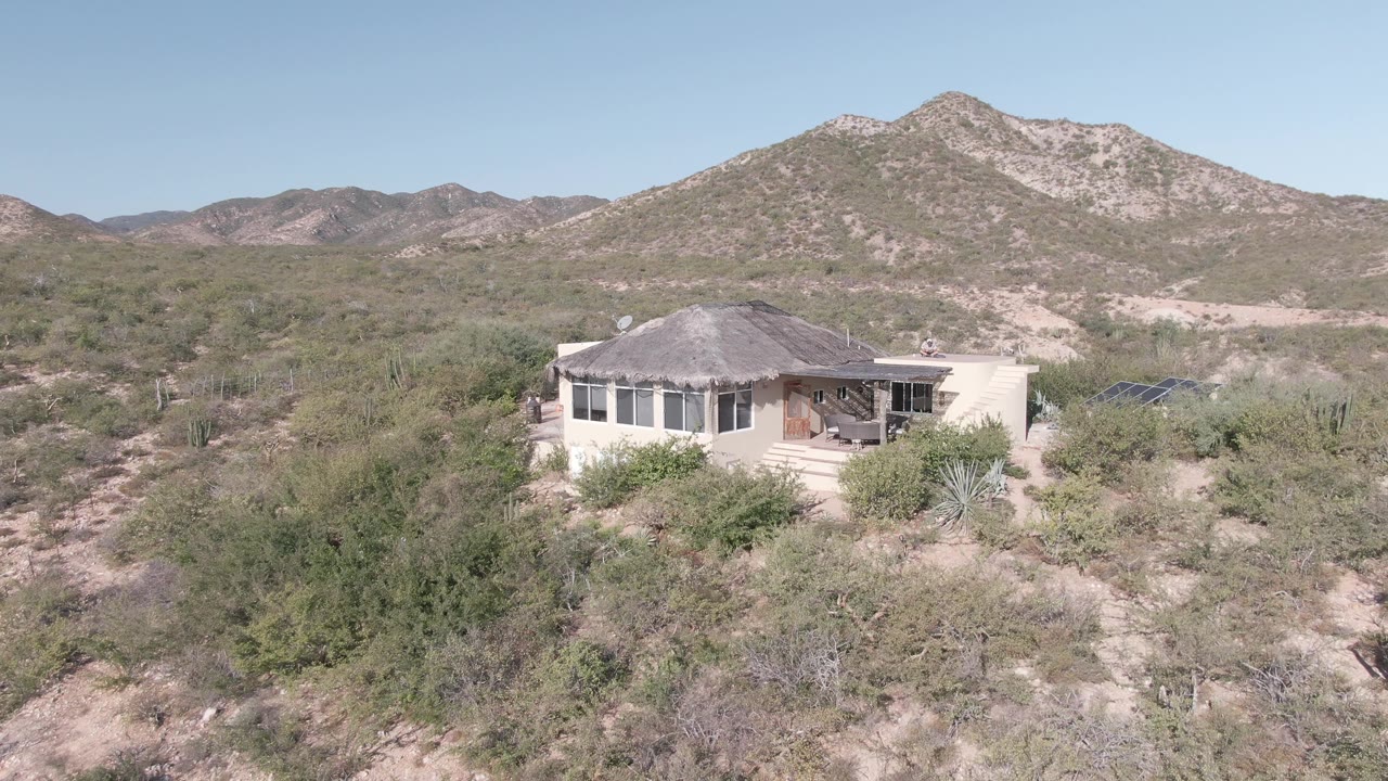 Cabo Pulmo desert residence seen from above with green brush in front, cactus around the structure, pale sandy tracks, mountain ridge, and no visible people.