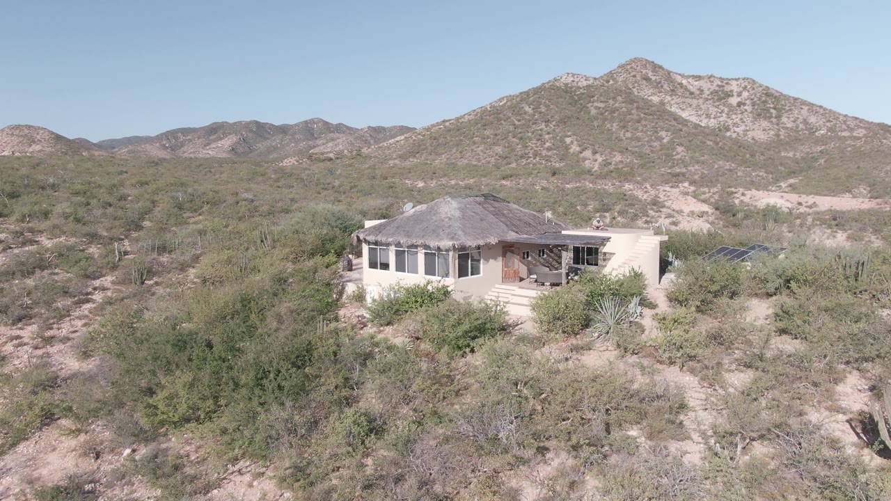 Light desert house in Cabo Pulmo, Baja California Sur, framed by dense scrub, visible cactus, pale dirt, distant hills, and no visible people.