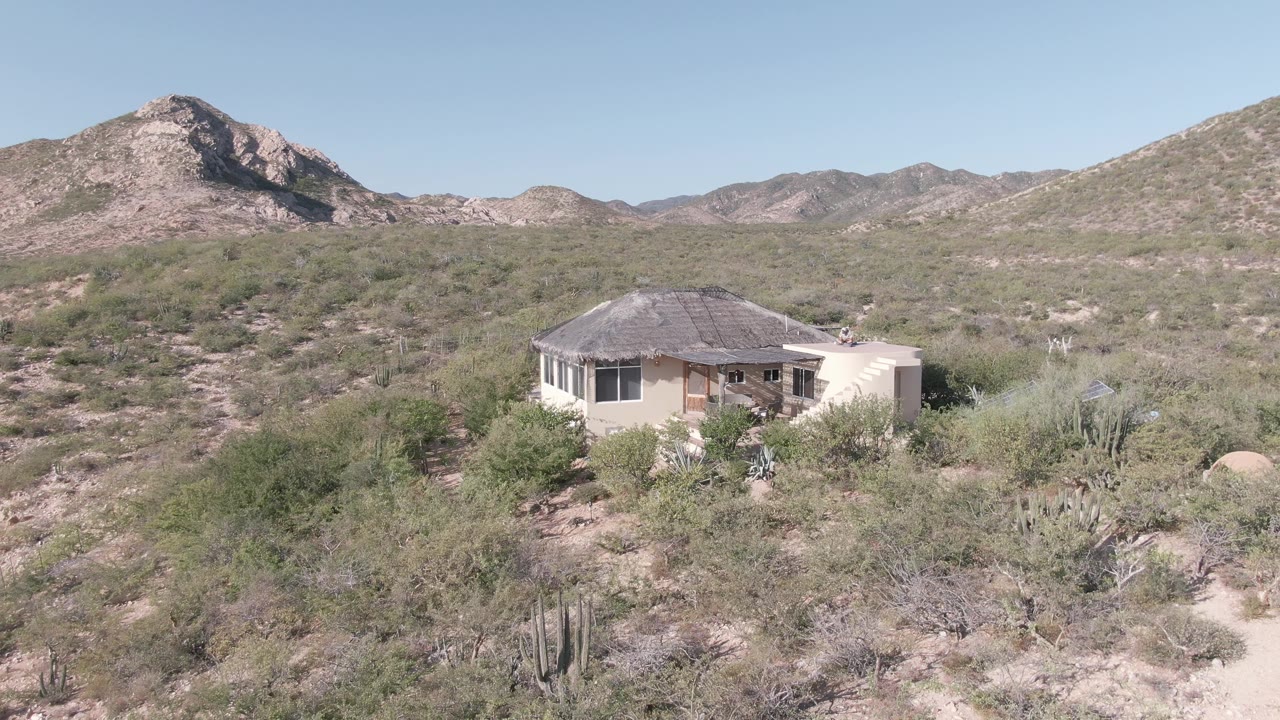 A small house in Cabo Pulmo, Baja California Sur sits among pale desert shrubs and cacti, with a rocky mountain behind it and no visible people.