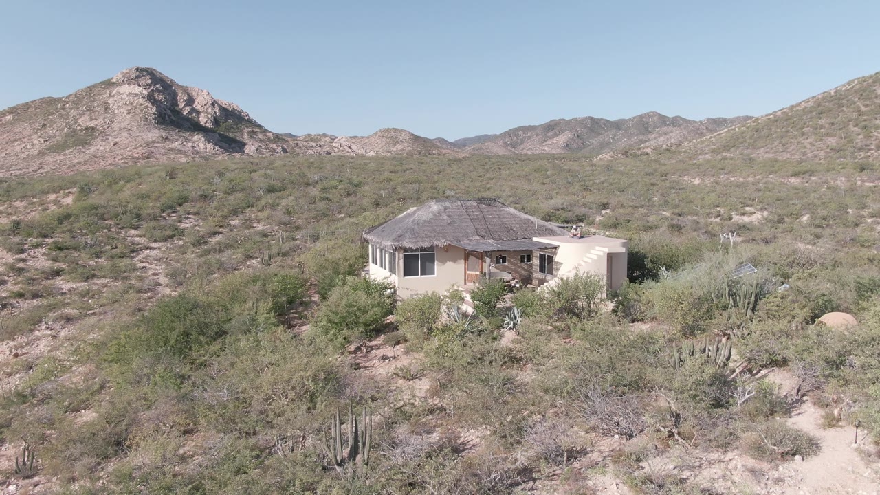 Remote desert house with cactus scrub and mountains under a clear sky.