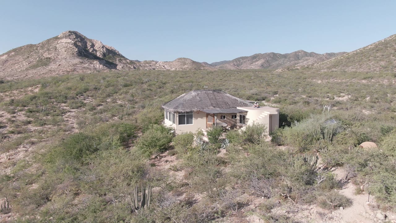A light-colored house in Cabo Pulmo stands amid dry brush and rocky ground, with a broad mountain ridge and clear sky behind it and no visible people.