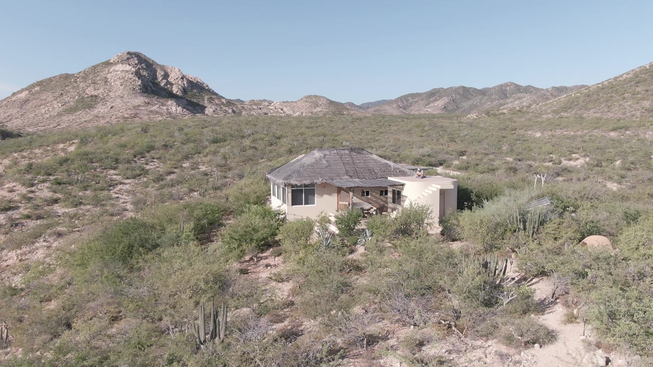 Desert home set back from cactus and shrubs with mountains in the distance.