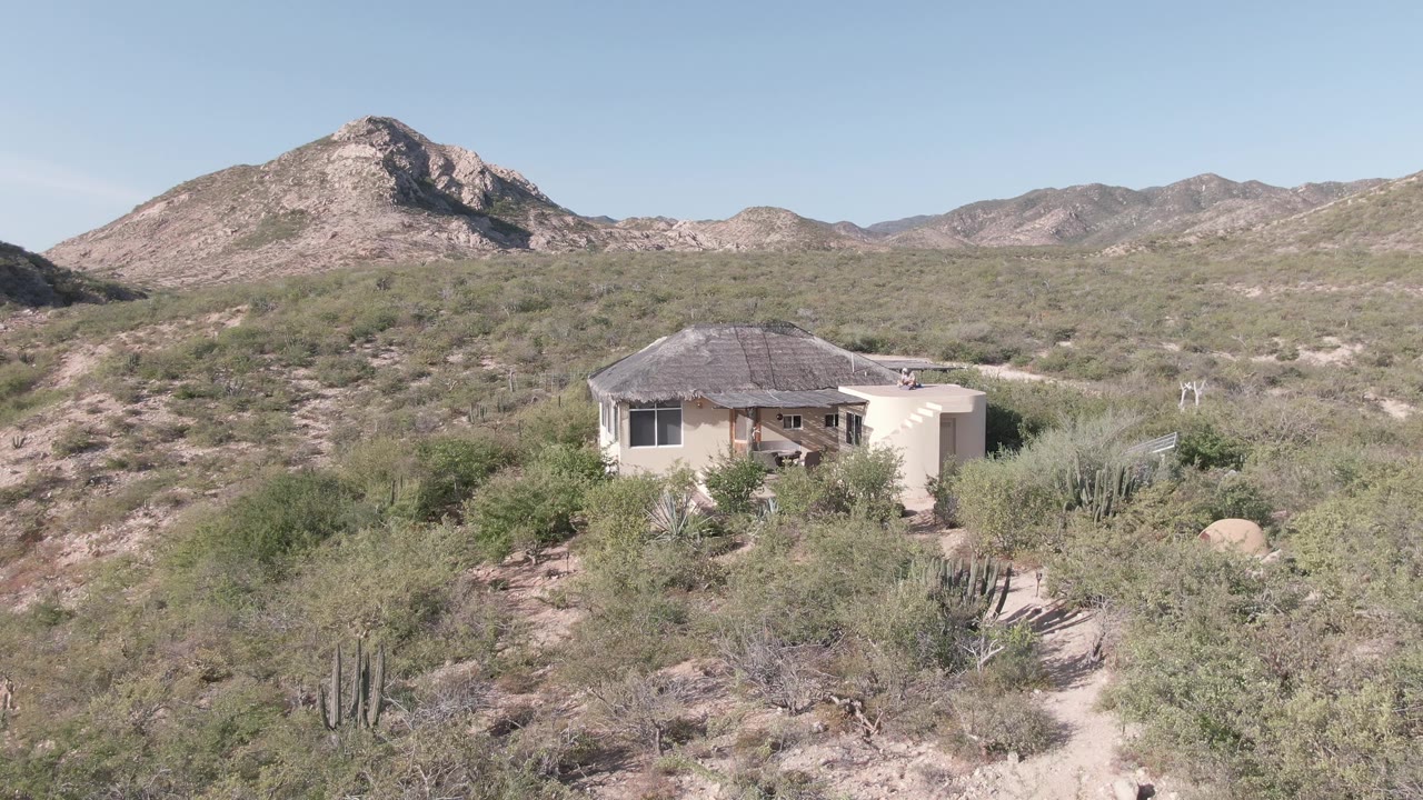 Wide elevated view in Cabo Pulmo, Baja California Sur, showing a small desert house among scrub and cacti below rocky mountains, with no visible people.