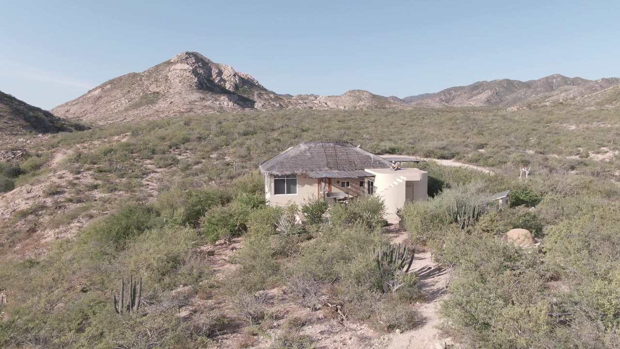 Elevated Cabo Pulmo view showing the house amid dense desert vegetation, a dirt path leading through the foreground, mountain ridges behind, and no visible people.