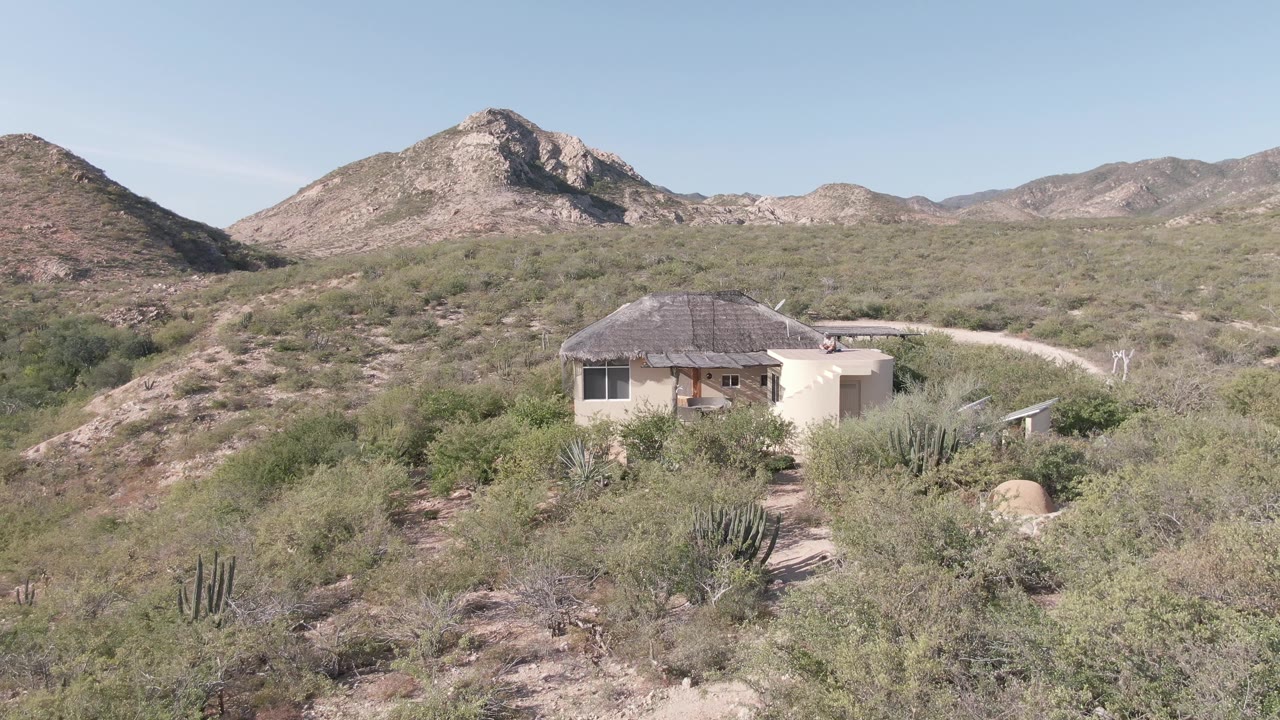 A small house in Cabo Pulmo sits low among cactus and desert shrubs, with rocky mountains behind it and no visible people in this opening frame.