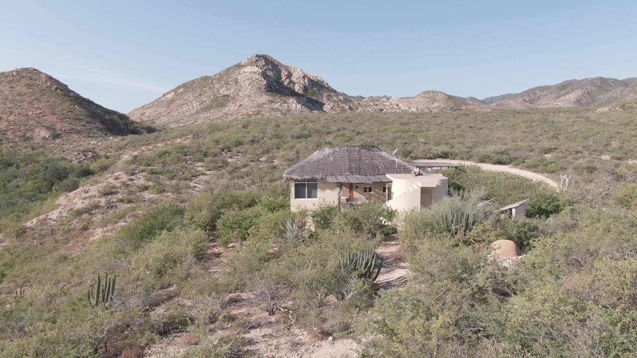 Desert house with porch, cactus, shrubs, dirt road, and rocky hills.