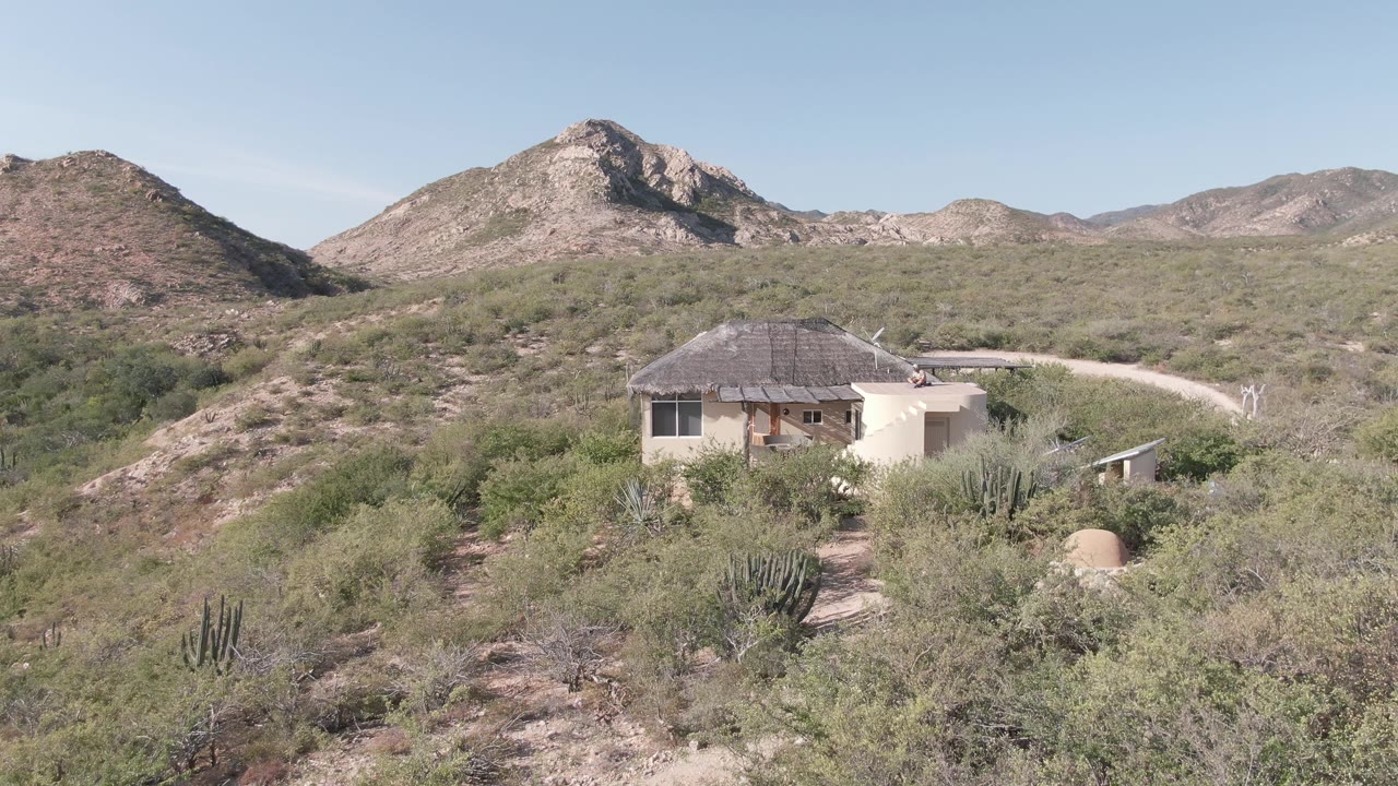 A pale house in Cabo Pulmo sits beneath the mountain ridge, with desert brush and cactus filling the foreground and no visible people in the frame.