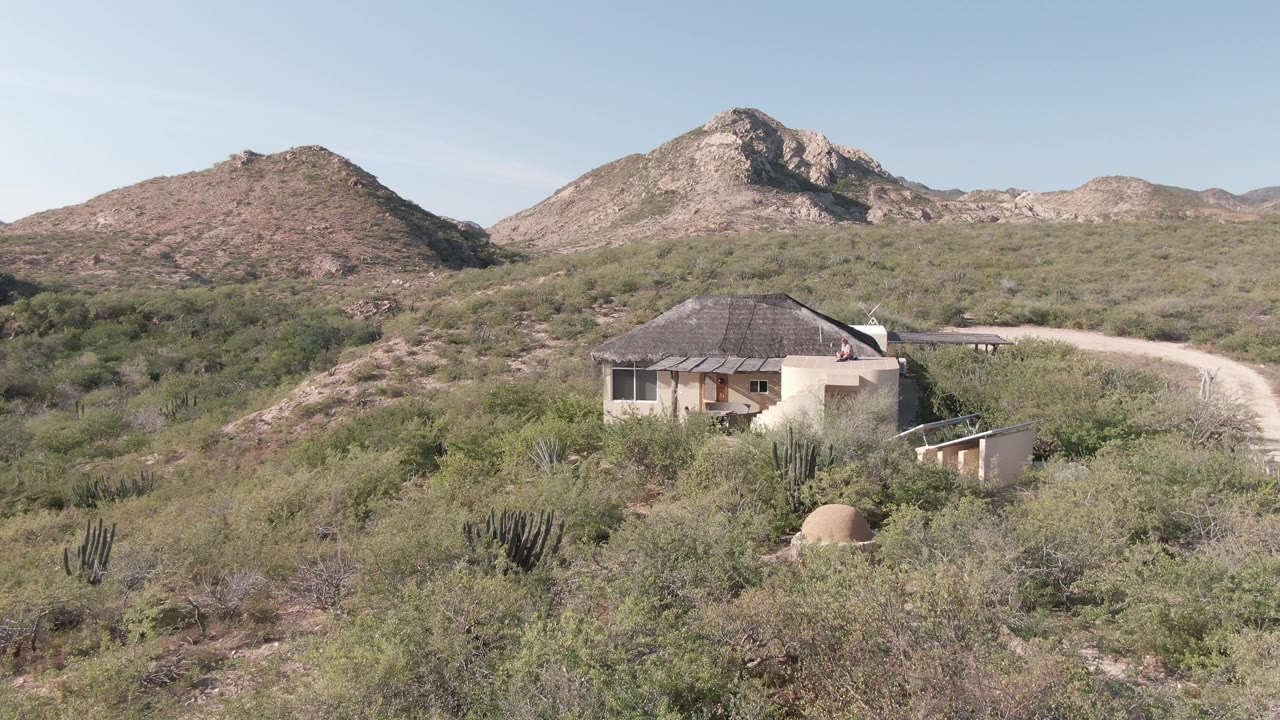 Wide view in Cabo Pulmo, Baja California Sur showing a pale cabin, upright cacti, green desert scrub, a sandy road, rocky hills, clear sky, and no visible people at the opening frame.