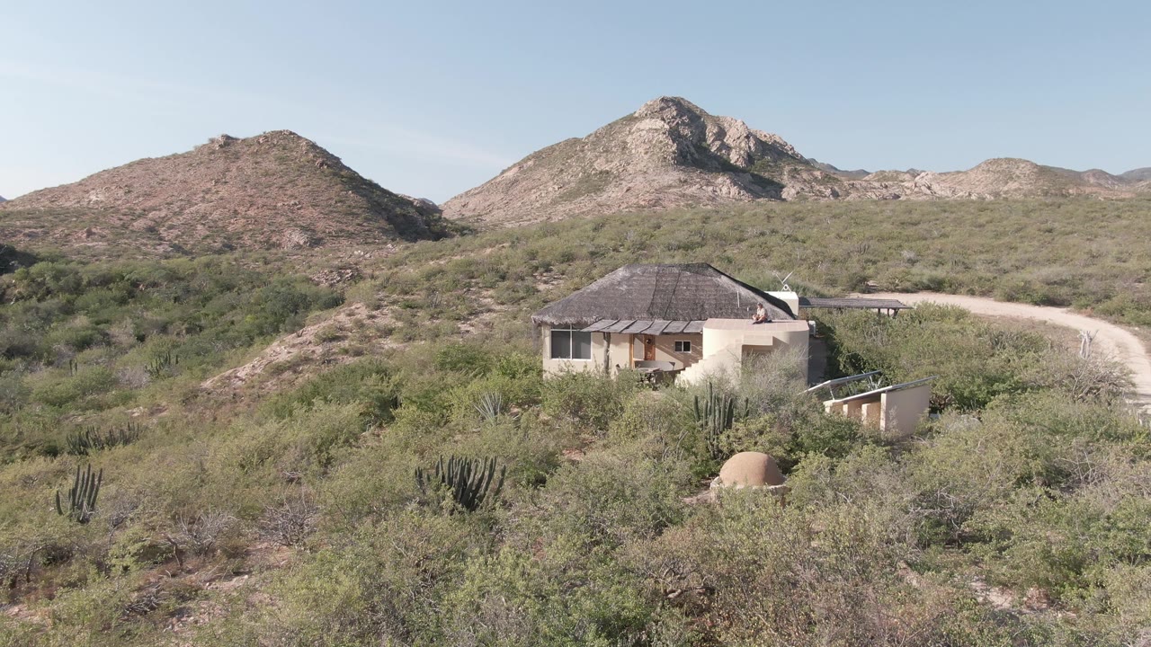 Desert cabin beside a sandy road with cacti and low mountains.