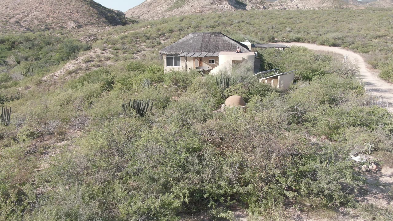 A pale rural residence in Cabo Pulmo appears among cactus and scrub, with the dirt road bending up the hillside and dry mountains visible across the background, with no visible people.