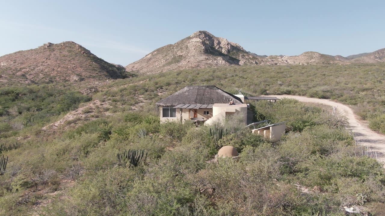 A pale house in Cabo Pulmo, Baja California Sur sits among desert brush and cacti near a curving dirt road, with low mountains in the background and no visible people.