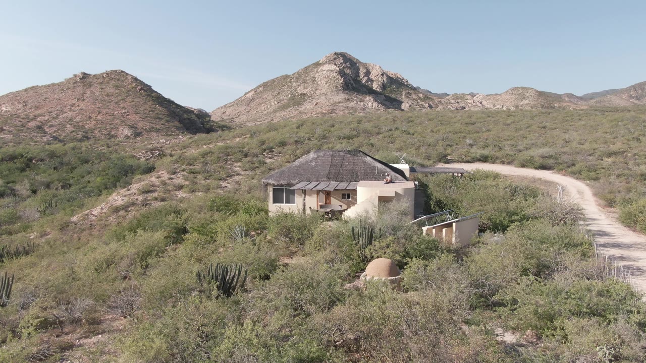 At one second, the Cabo Pulmo desert house remains centered below the hills, with green scrub in the foreground, the dirt road on the right, and no visible people.
