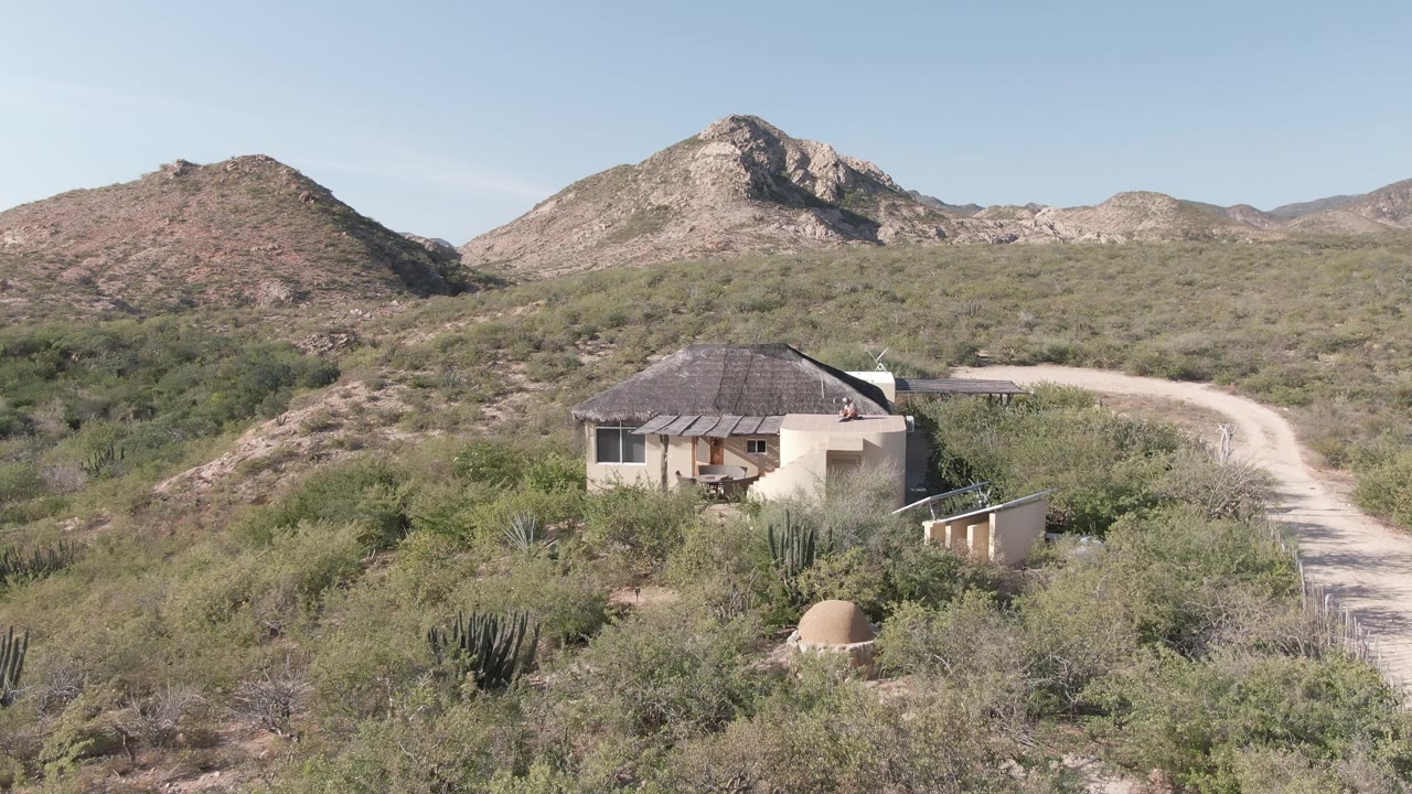 At two seconds, the elevated view shows the pale house partly surrounded by vegetation, a bright dirt road to the right, rocky Cabo Pulmo hills, and no visible people.