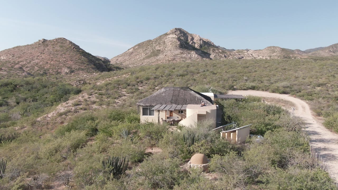 Desert house below rocky hills with brush and a dirt road nearby.