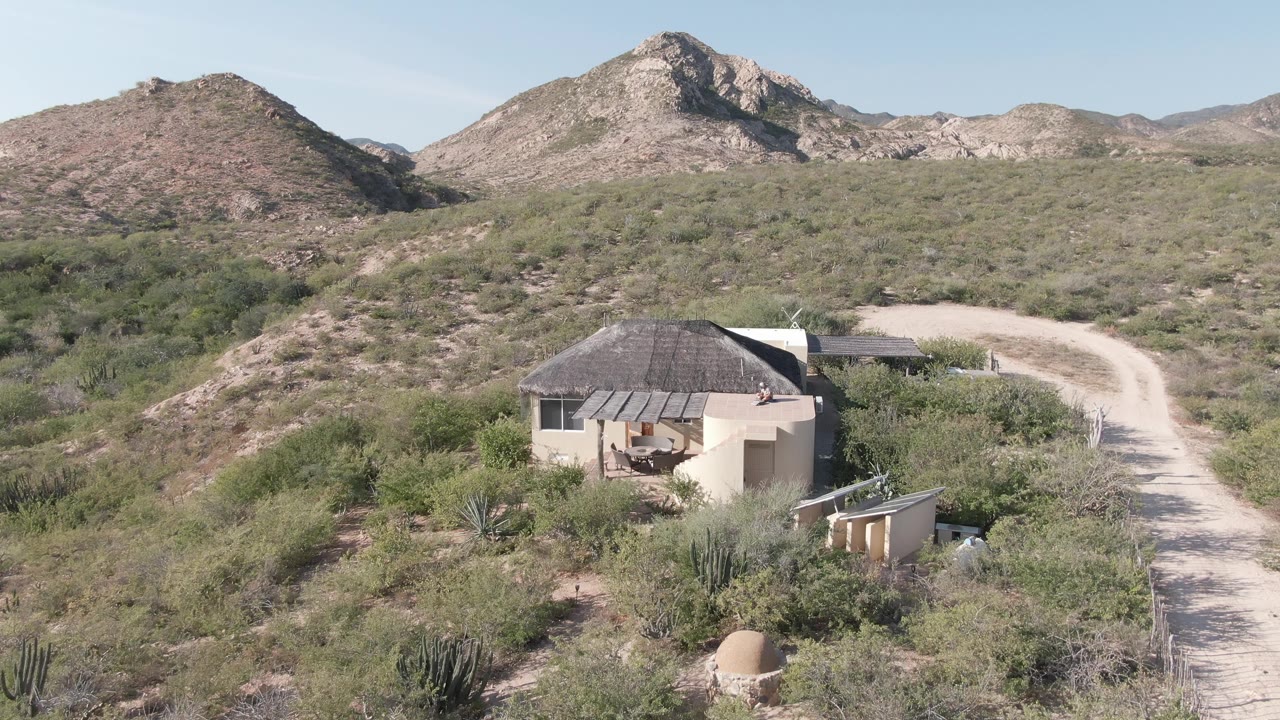 Aerial frame from Cabo Pulmo, Baja California Sur showing a small round hut, scrubby desert vegetation, a curving pale dirt road, rocky hills, clear sky, and no visible people.
