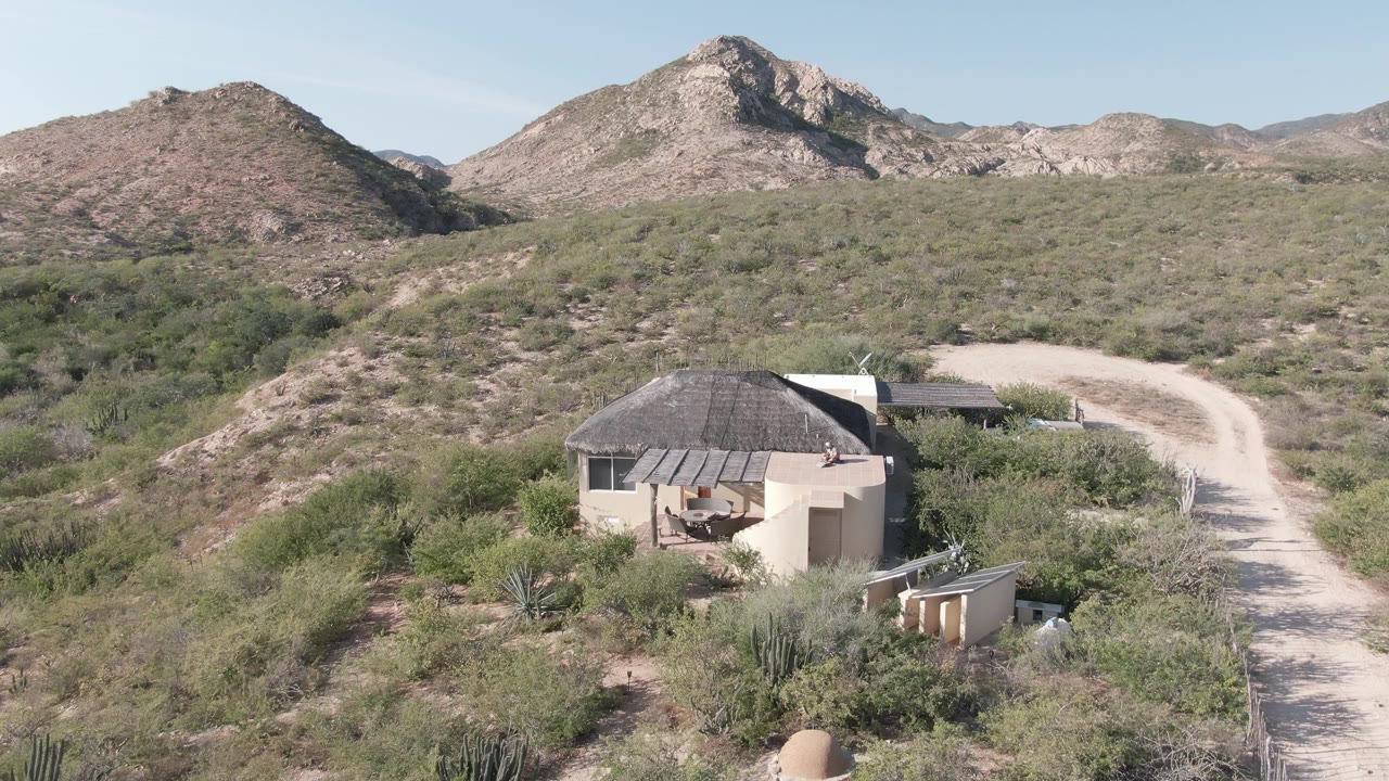 Aerial view in Cabo Pulmo, Baja California Sur with the small hut lower left, the dirt road curving up the right side, dense desert shrubs, distant rocky hills, and no visible people.