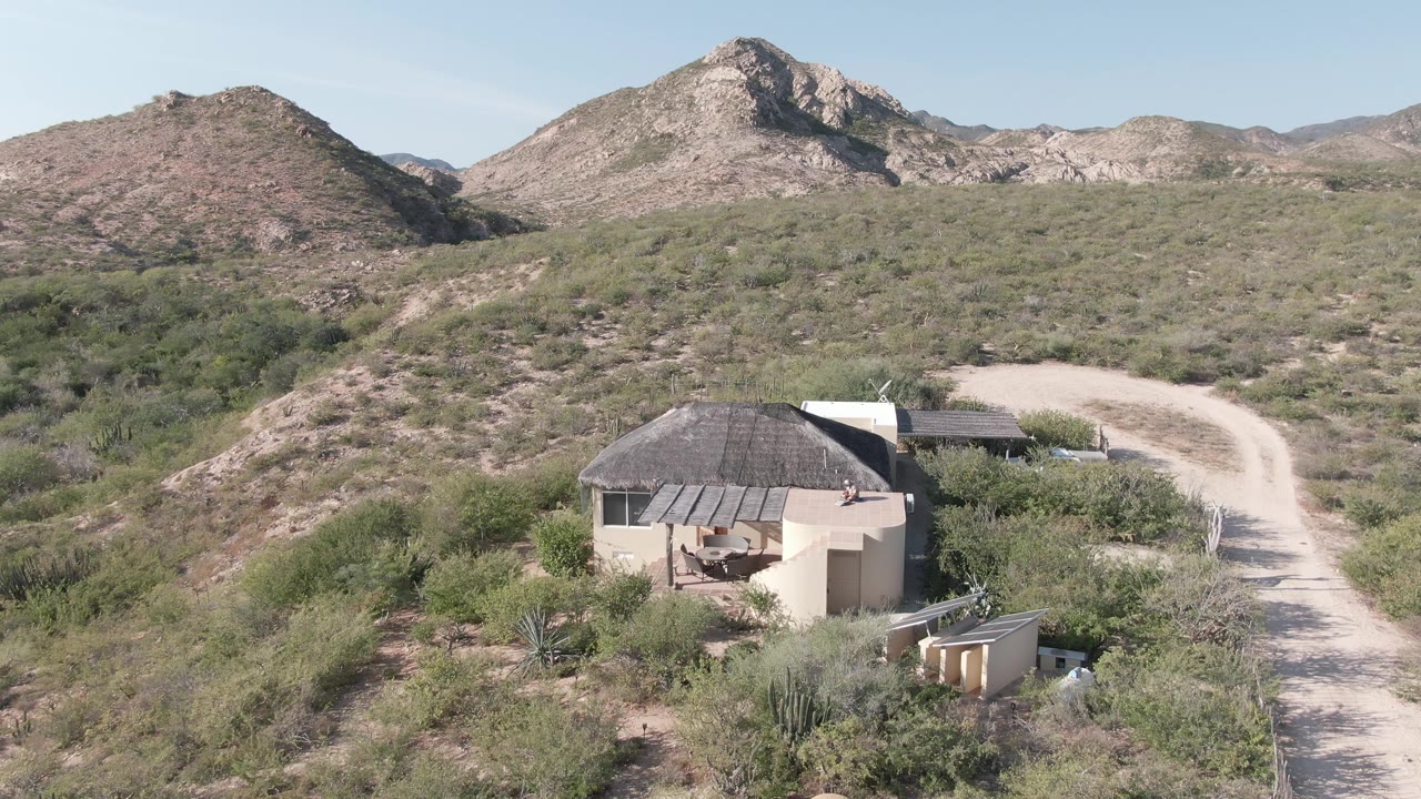 Cabo Pulmo aerial frame showing the hut slightly left of center, a pale road sweeping around the right hillside, scattered cacti and shrubs, dry mountains, clear sky, and no visible people.
