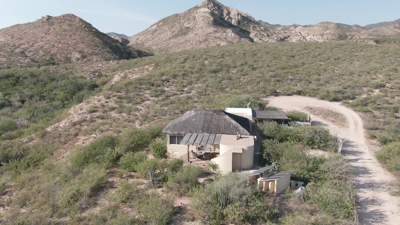 Wide aerial of a hut below a hillside road in a dry mountain landscape.