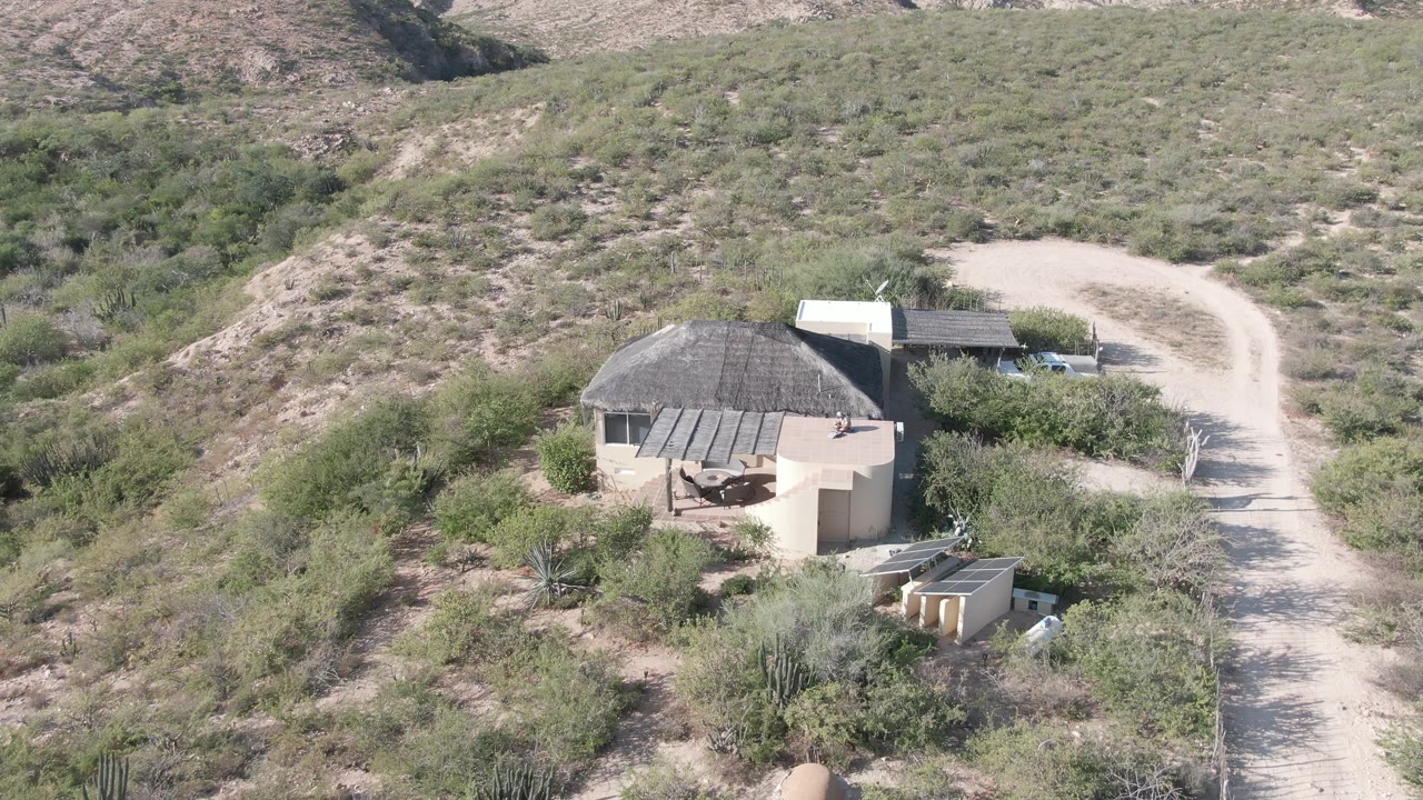 Aerial frame in Cabo Pulmo, Baja California Sur showing a round tan structure beside a curving dirt road, sparse desert shrubs, low hills, and no visible people.