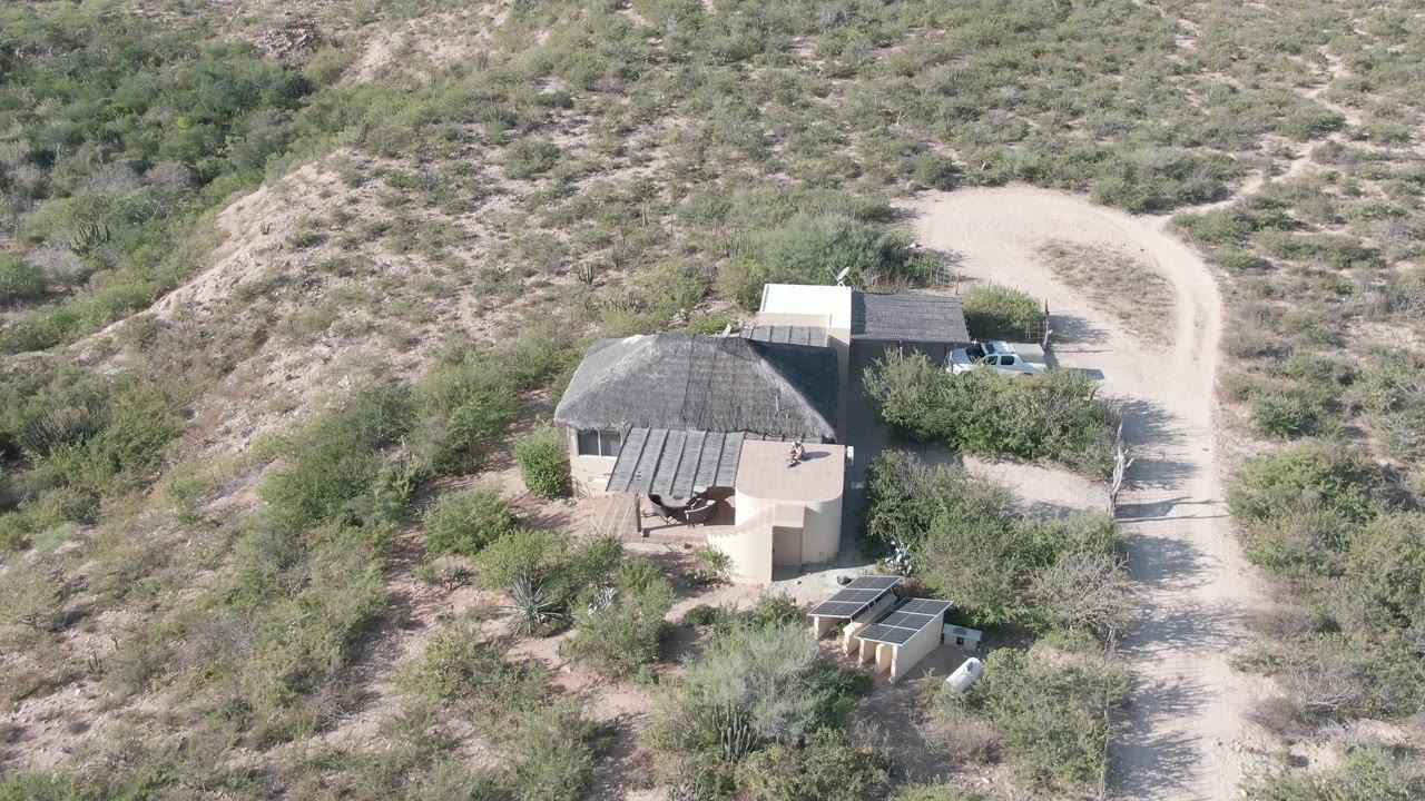 Aerial view in Cabo Pulmo showing the thatched-roof house left of a pale dirt road, with the white truck still near the roadside and no visible people.