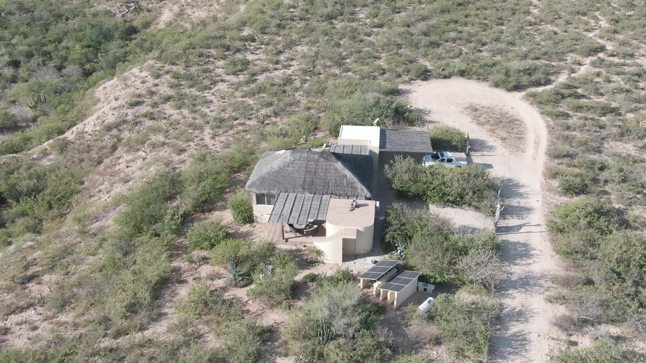 Drone-height frame in Cabo Pulmo with the house, adjacent outbuildings, white truck, curving dirt road, dry hillside, and no visible people.