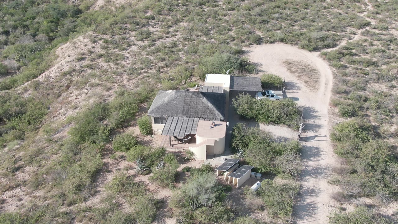 Aerial shot of a desert house below a scrub-covered slope and beside a dirt road.