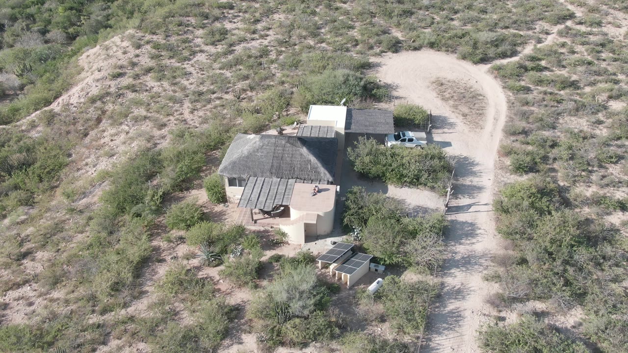 Aerial frame over a rural desert house in Cabo Pulmo, Baja California Sur, with small structures, a pale dirt road, scrub-covered hills, a white vehicle near the property, and no visible people.