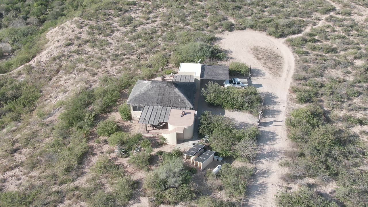Aerial view in Cabo Pulmo, Baja California Sur, looking down at a tan desert house, nearby outbuildings, scattered trees, a curving dirt road, dry hillside vegetation, and no visible people.