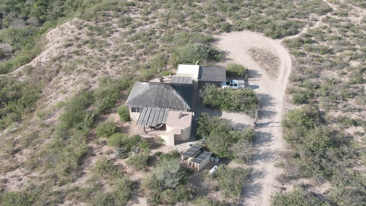 Overhead frame of the Cabo Pulmo desert property with the house in the lower portion, a light dirt road bending around the right side, dry brush across the hillside, and no visible people.