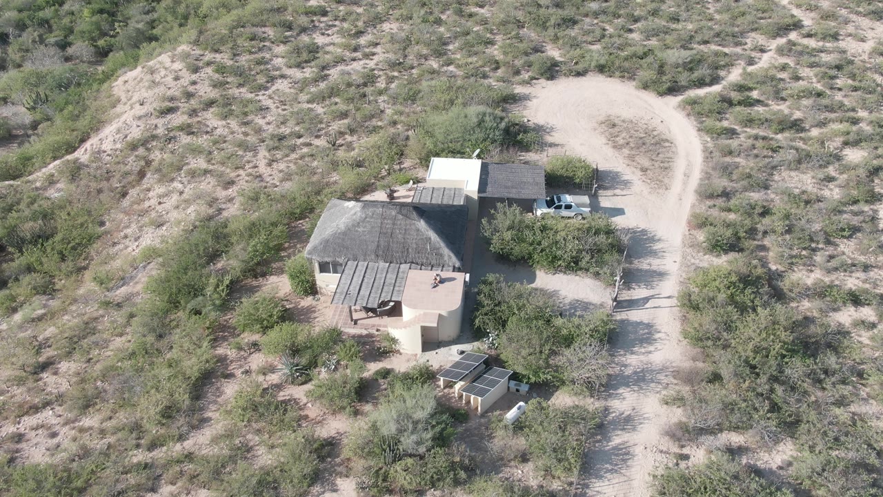 Desert house and small structures viewed from above beside a dusty road.
