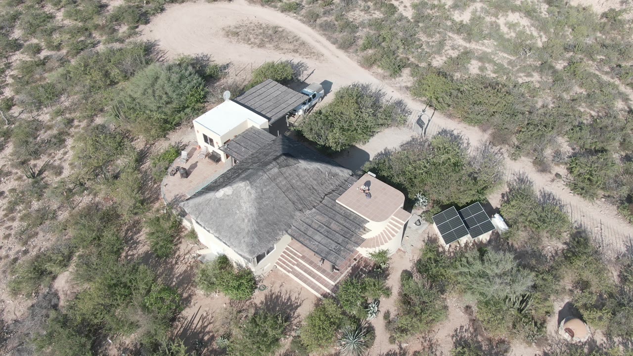 High aerial frame over a Cabo Pulmo desert home, showing a pale house, dark roof, solar panels, sandy road, scrub vegetation, and a small person on the roof near the start of the clip.