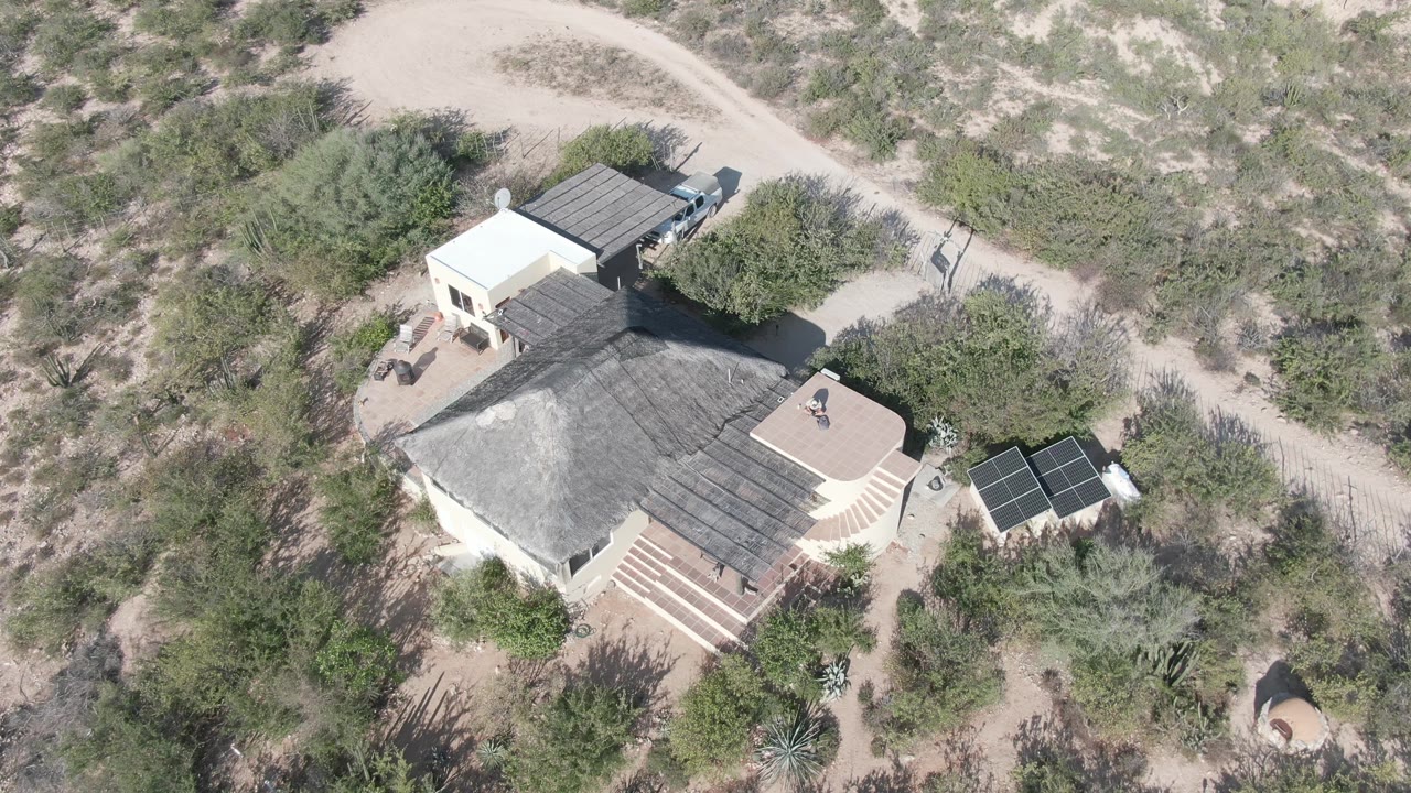 Drone-style overhead angle on the Cabo Pulmo residence, with the small figure still on the roof, solar panels beside the home, and dry brush filling the surrounding sandy lot.