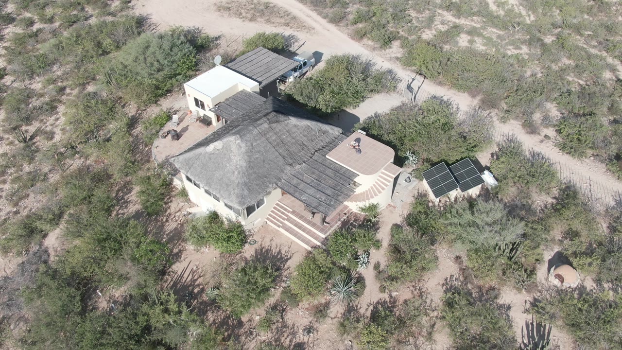 High aerial frame over a pale desert house in Cabo Pulmo, Baja California Sur, with solar panels, sandy tracks, cactus vegetation, and a tiny person visible on the roof.