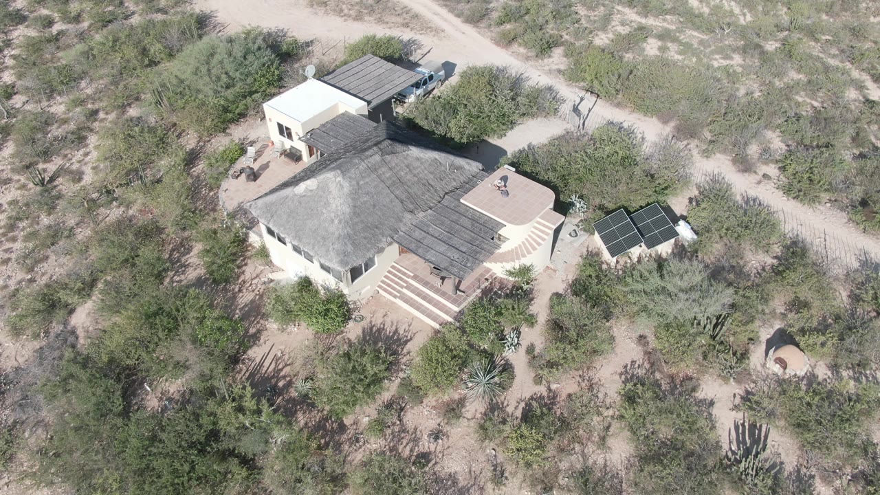 Aerial frame over the Cabo Pulmo desert house with the roof, entry steps, solar panels, sandy road, and surrounding cactus vegetation visible, with no visible people.