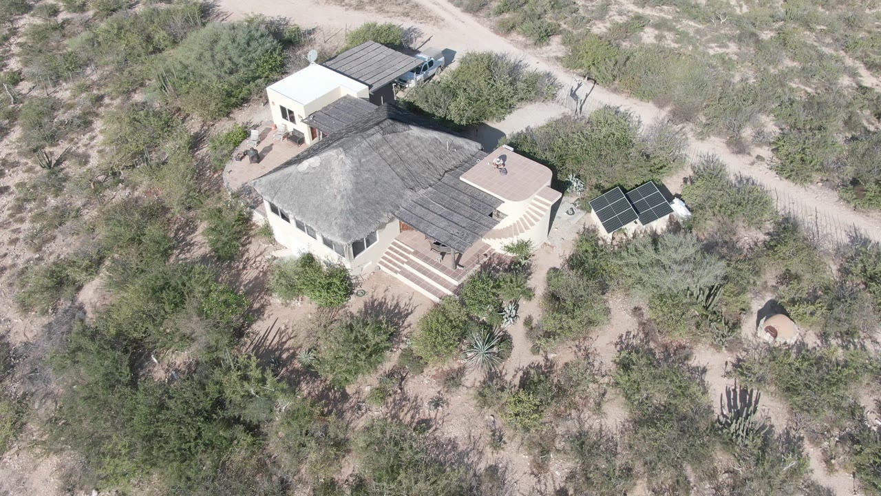 High drone-style view of the Cabo Pulmo desert property, showing the pale house, dark roof, solar panels to the right, dirt roads above it, and no visible people.