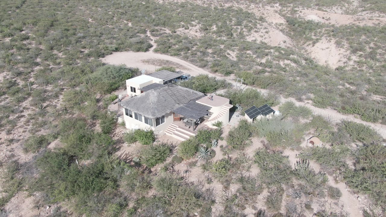 Aerial view in Cabo Pulmo, Baja California Sur, showing a pale house with a gray roof, steps, solar panels, sandy tracks, scrub vegetation, and no visible people.
