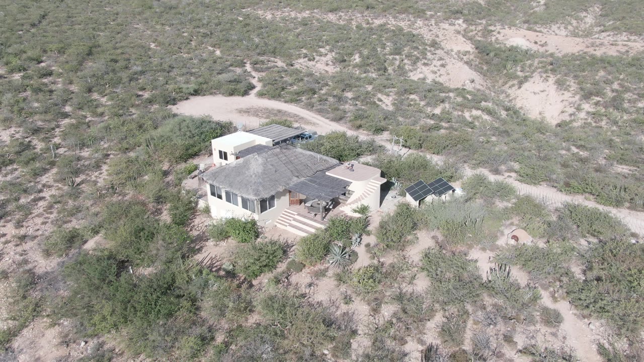 Elevated Cabo Pulmo frame with the house slightly left of center, solar panels to the right, dry shrubs around the structure, pale hillside terrain behind it, and no visible people.