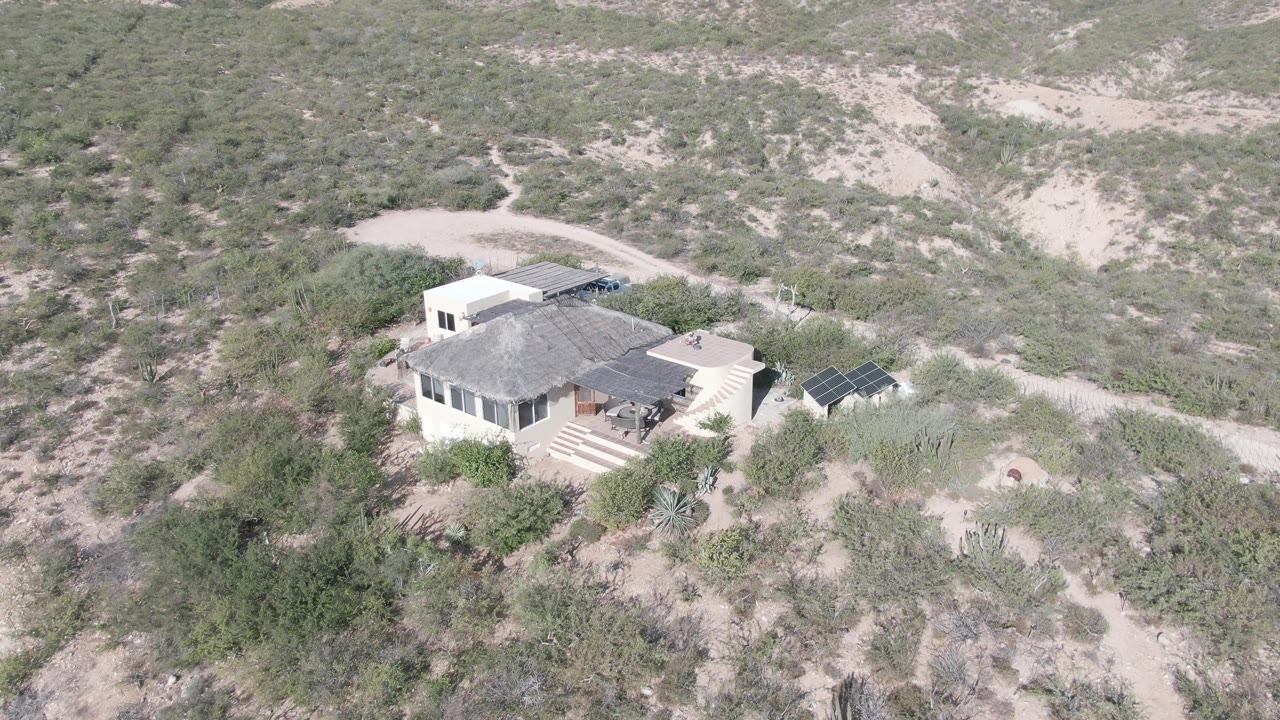 Cabo Pulmo aerial view with the pale house lower in frame, a dark solar-panel array on the right, sandy paths cutting through green scrub, and no visible people.
