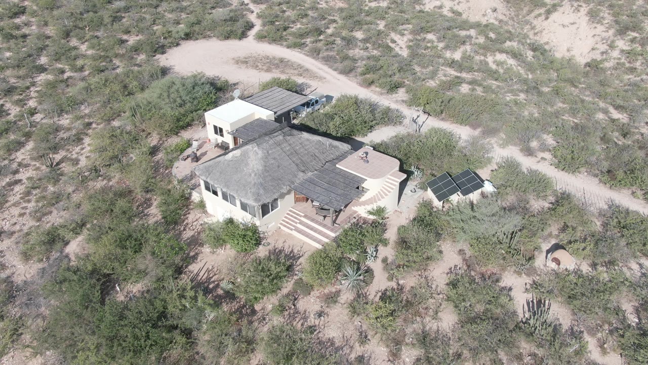 Aerial frame over a pale house in Cabo Pulmo, Baja California Sur, with a dirt track behind it, solar panels to the right, and dry scrub around the property.