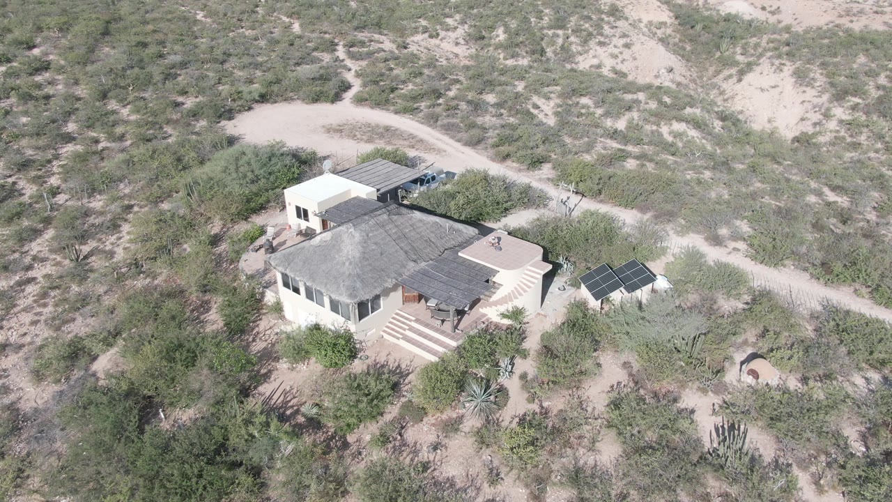 A high aerial frame in Cabo Pulmo, Baja California Sur, places the house below the sandy hillside, with the solar panels clearly visible at right and no close facial detail visible.