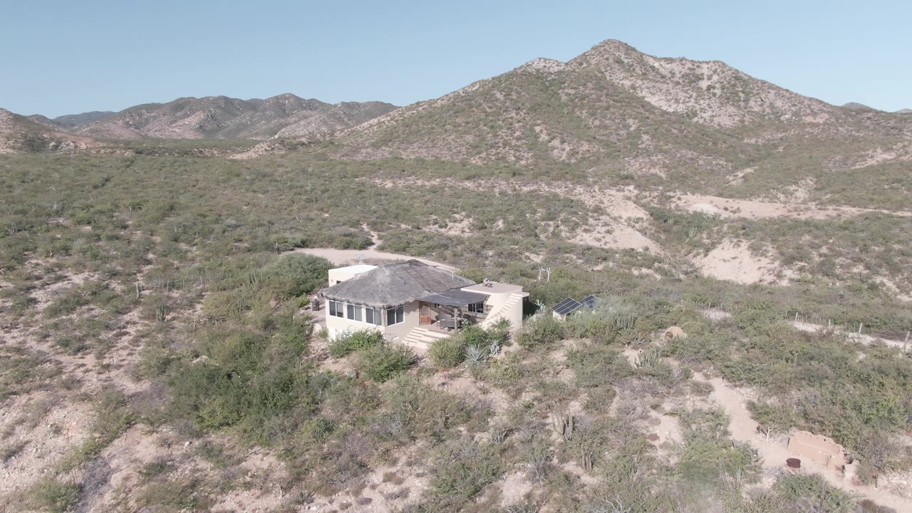 Aerial frame from Cabo Pulmo, Baja California Sur showing a pale house low in the left foreground, sandy desert washes, green shrubs, rocky mountains, clear sky, and no visible people.