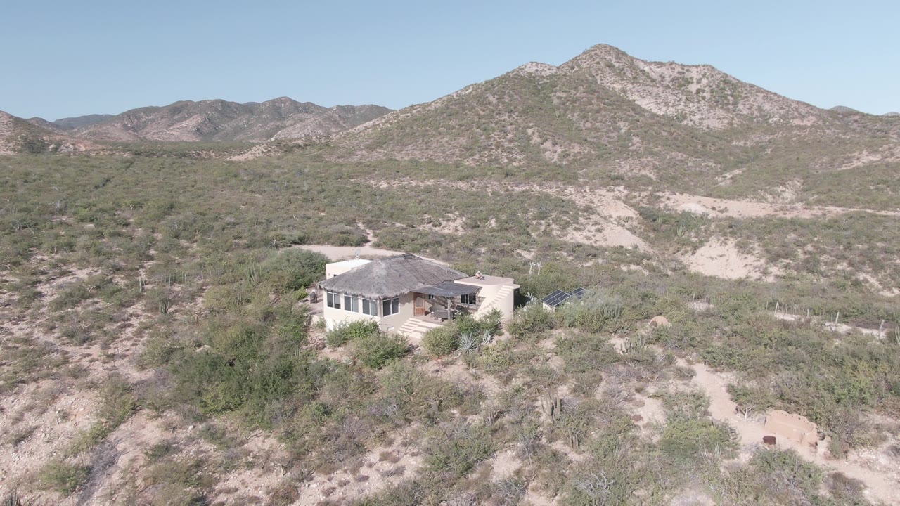 Cabo Pulmo aerial view with the house slightly nearer the lower center, scrub-covered desert slopes, pale sandy tracks, distant rocky hills, clear sky, and no visible people.