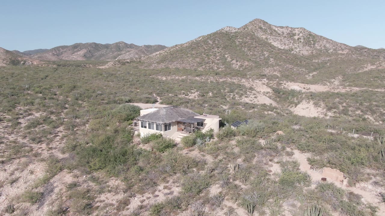 Elevated Cabo Pulmo frame showing the house amid dense low shrubs, cactus-like vegetation near the foreground, pale sandy cuts in the terrain, mountain ridges, bright daylight, and no visible people.